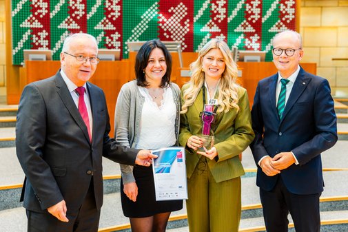 Zwei Frauen und zwei Männer nebeneinander. Die Frau in der Bildmitte hat blonde, lange Haare und trägt einen grünen Hosenanzug, in der Hand den vilmA-Preis.