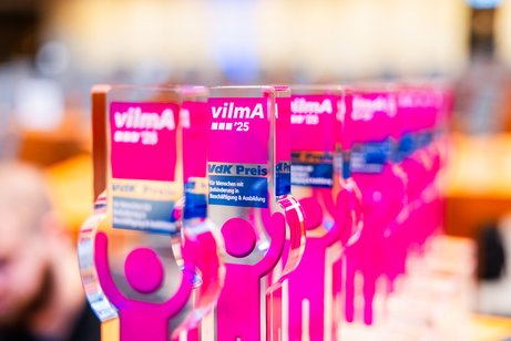 Acht vilmA-Pokale stehen hintereinander - pinke Icons in Menschenform, die ein Schild hochhalten mit weißer Schrift: vilmA '25.