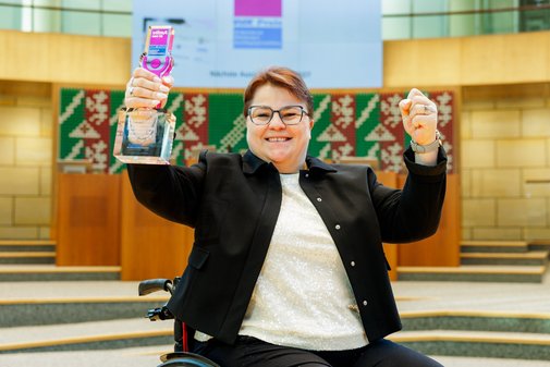 Eine Frau im Rollstuhl mit dunkler Kurzhaarfrisur und Brille hält den vilmA-Preis in der linken Hand, die rechte ist ebenfalls emporgereckt zur Siegerfaust.