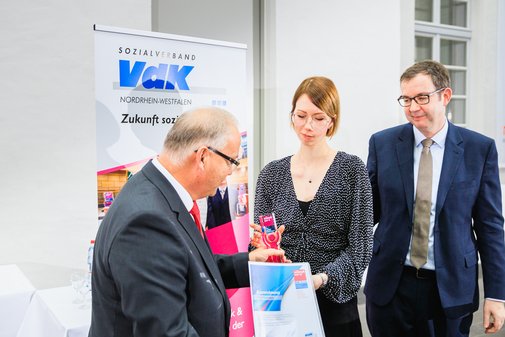 VdK-Vorstand Thomas Zander gemeinsam mit einer Preisträgerin und einem Laudatoren auf der Bühne vor VdK-vilmA-Banner.