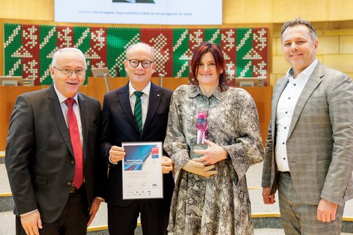 Vier Menschen stehen nebeneinander: Zwei Männer in dunklem Anzug, einer hält eine vilmA-Urkunde, eine Frau in geblümten Kleid mit vilmA-Preis in der Hand, rechts von ihr ein Herr in hellem Anzug,