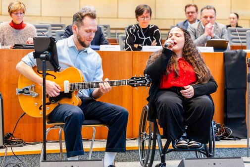 Ein junger Mann mit dunkelblonden Haaren und Bart sitzt Gitarre spielend auf einem Stuhl. Neben ihm im Rollstuhl eine junge Frau mit langen lockigen Haaren, die in ein Mikrofon singt.