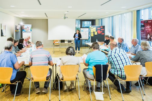 Menschen sitzen in einem Seminarraum an u-förmig aufgestellten Tischen. Vor Kopf steht eine Referentin vor einem White-Board.